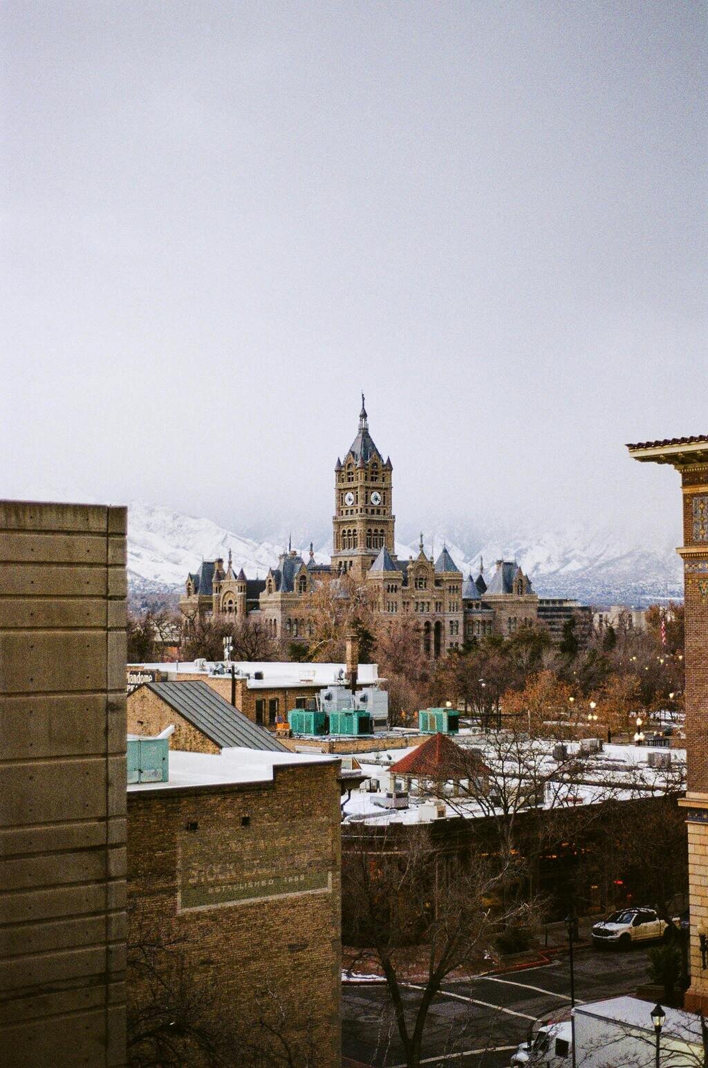 A historic clock tower building surrounded by trees and snow-covered rooftops, with mountains in the background under a cloudy sky.