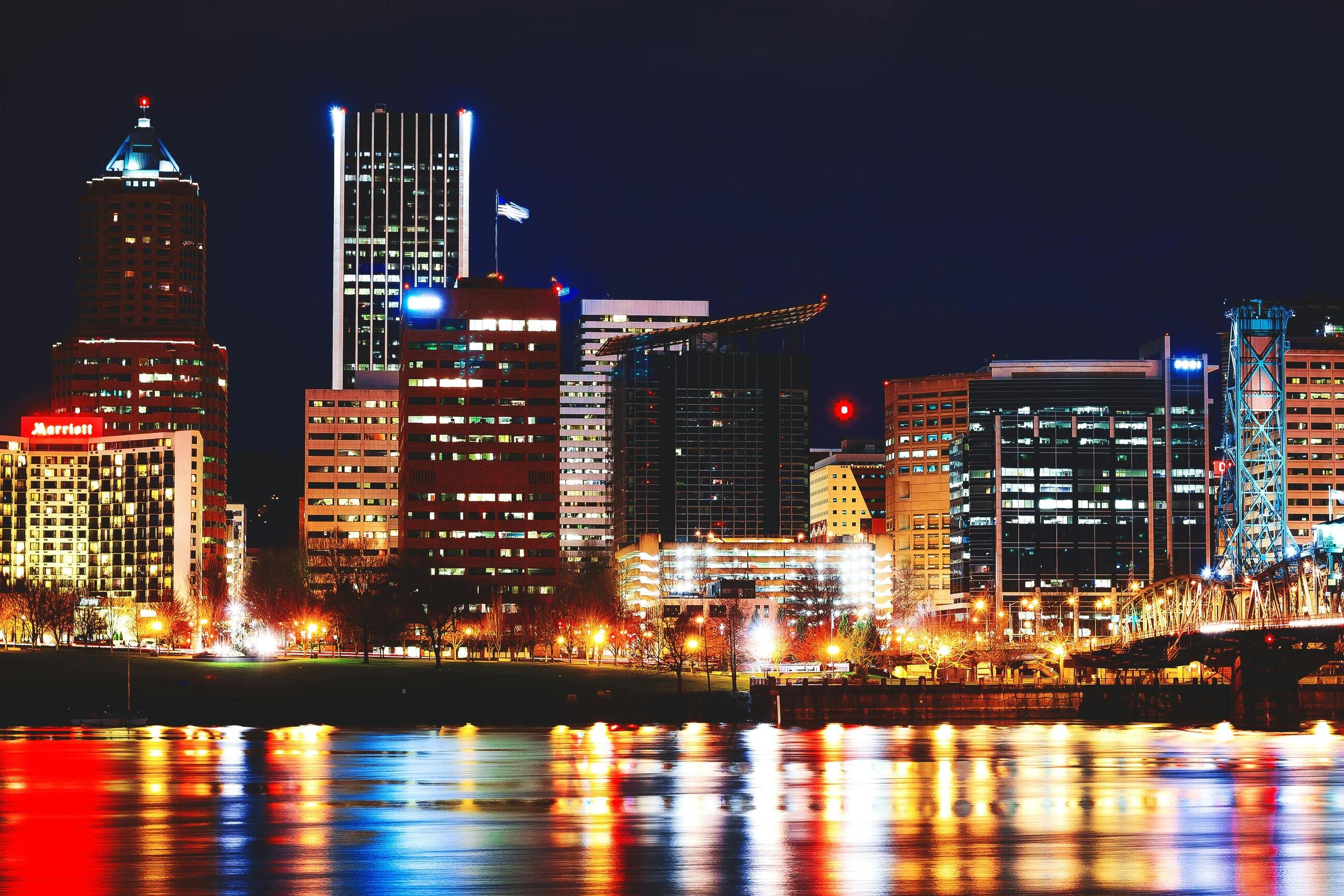 A vibrant city skyline at night with illuminated buildings, colorful reflections on the water, and a lit bridge on the right.