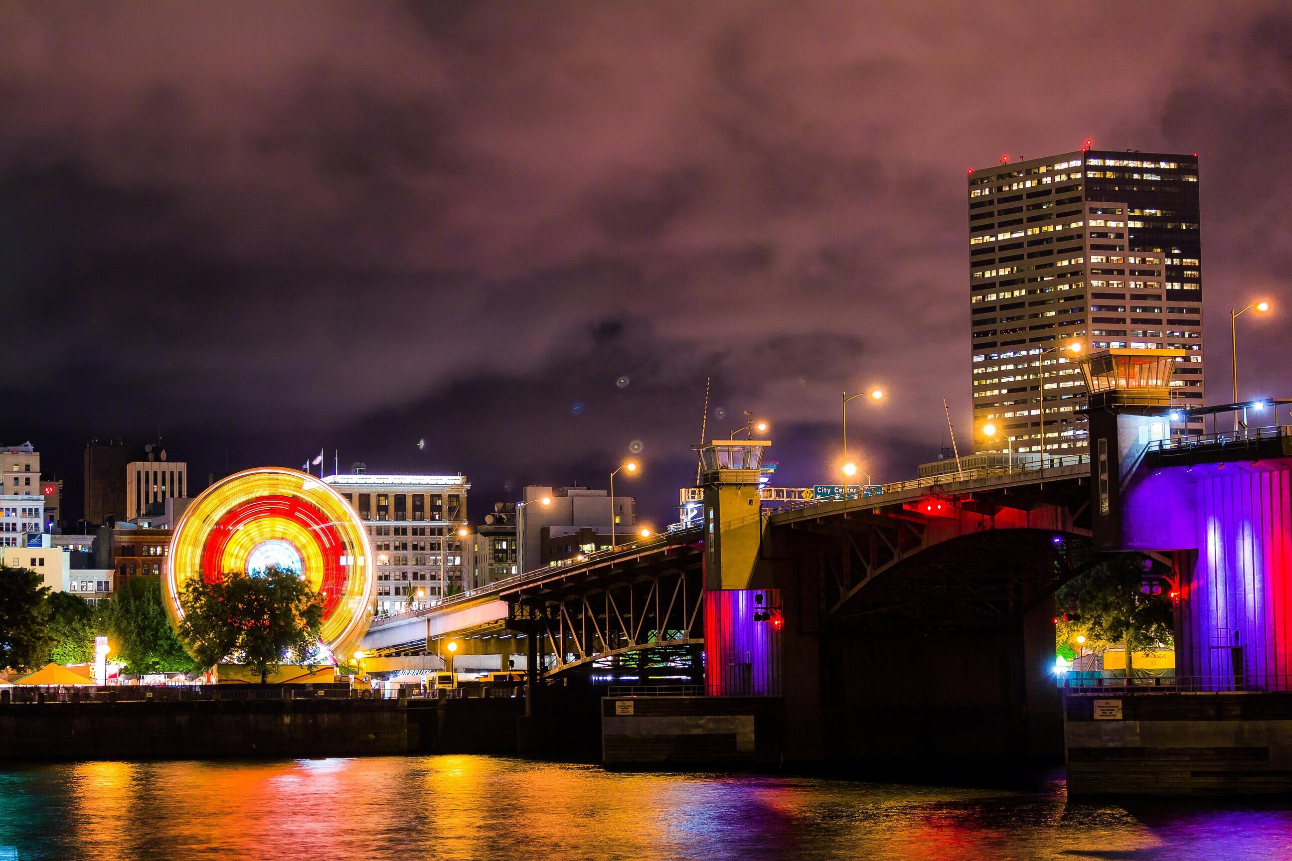 A brightly lit Ferris wheel and a bridge at night, with city buildings and colorful reflections on the water below.