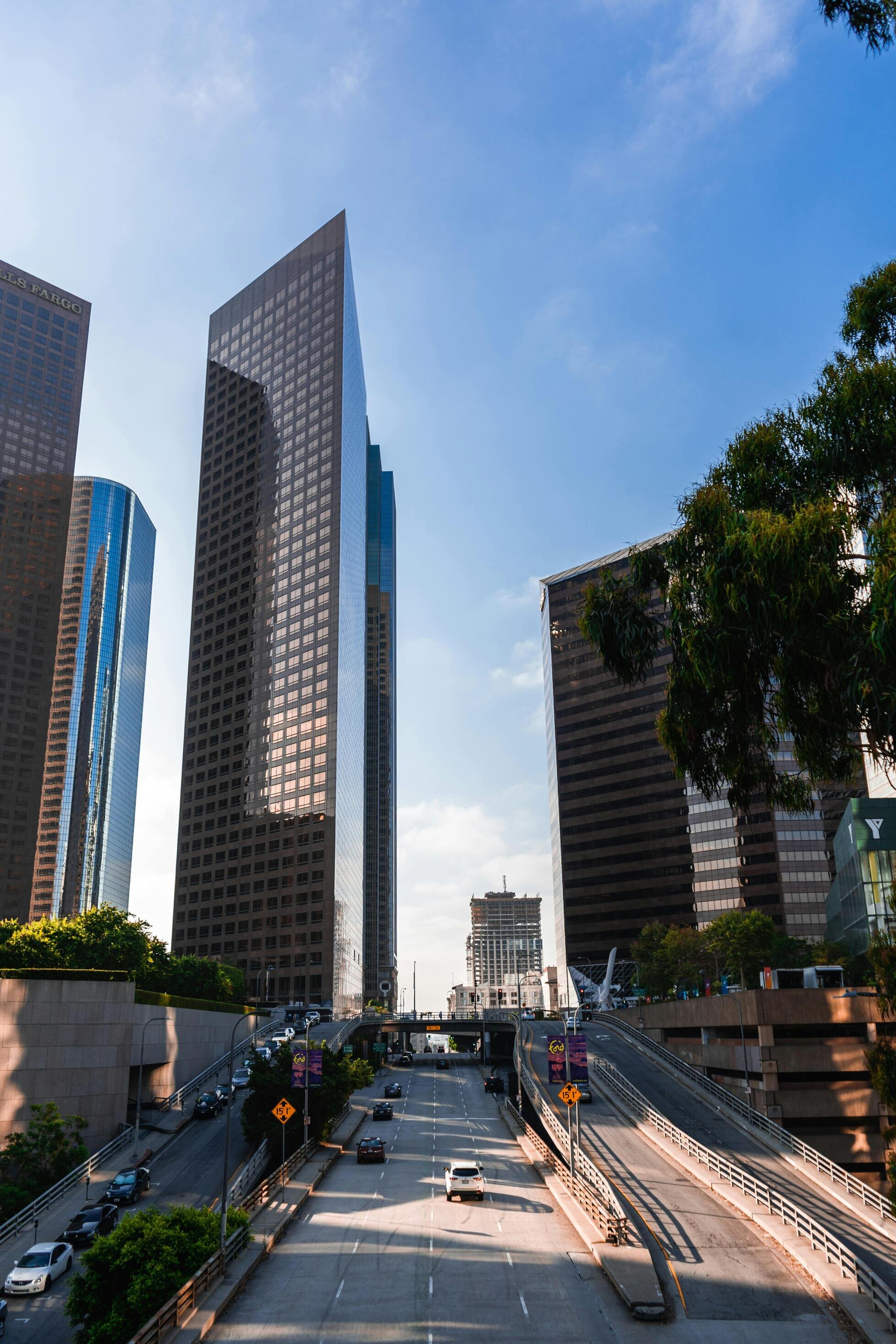 A city street surrounded by tall glass skyscrapers under a clear blue sky, with cars driving through the urban setting.