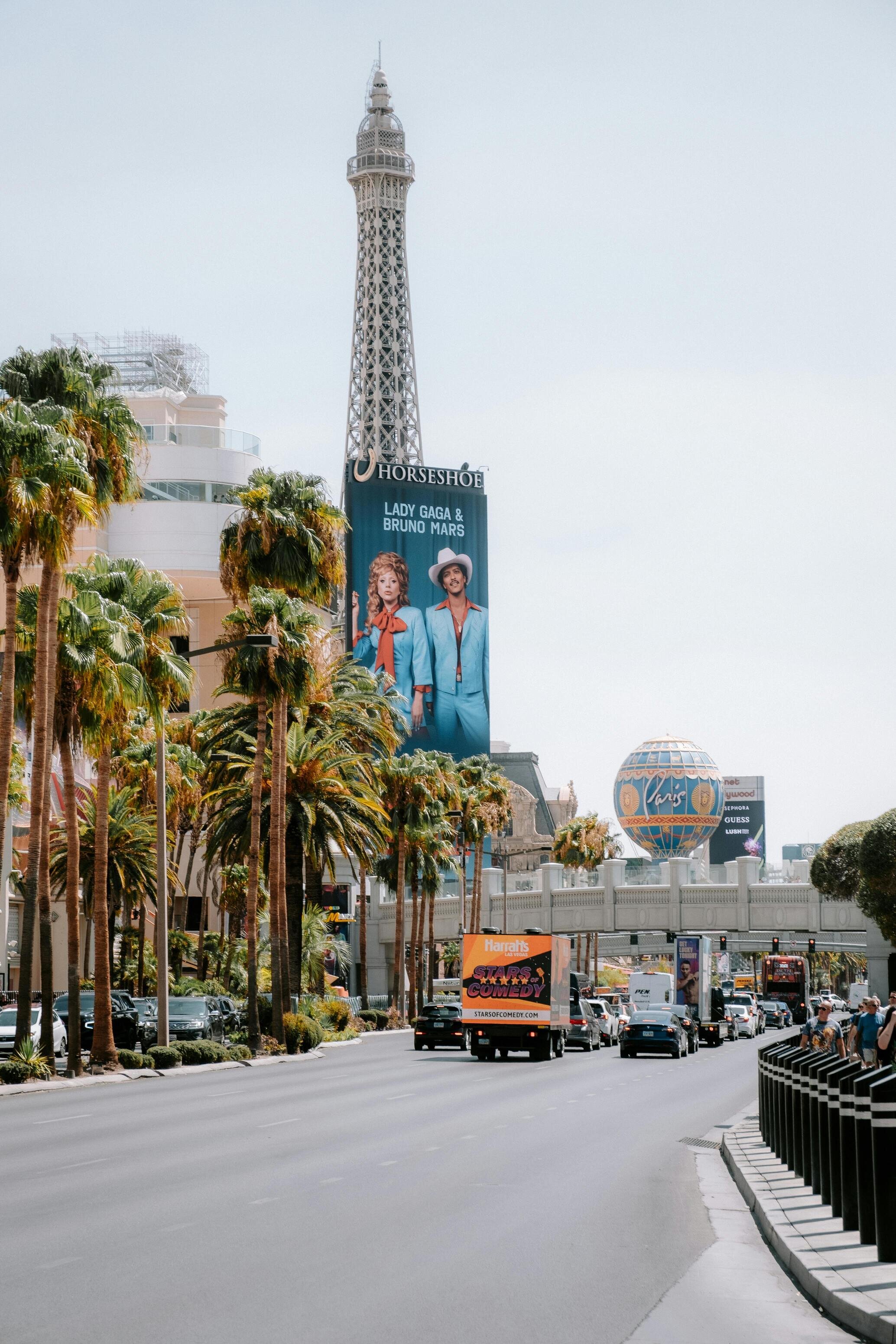 A busy city street lined with palm trees, featuring a replica of the Eiffel Tower and vibrant billboards, including one promoting Lady Gaga and Bruno Mars.