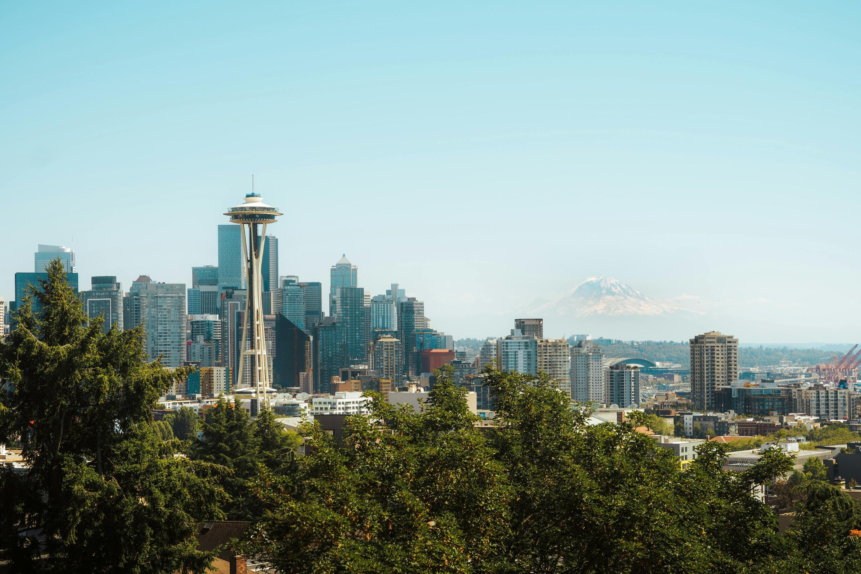 A city skyline featuring the Space Needle, modern buildings, and Mount Rainier in the background, with trees in the foreground.