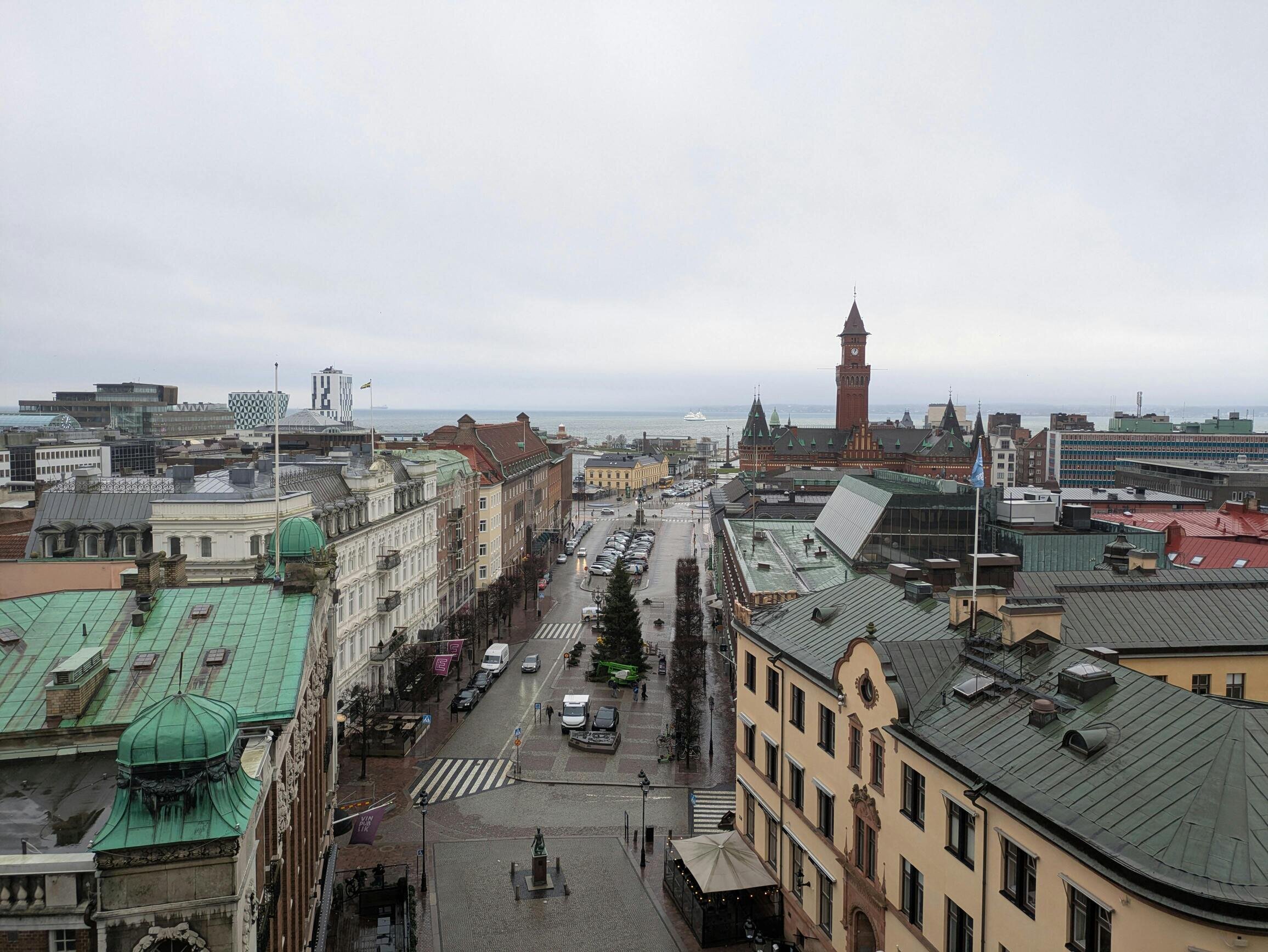 A historic cityscape with old buildings featuring green rooftops, a clock tower, and a view of the sea under a cloudy sky.