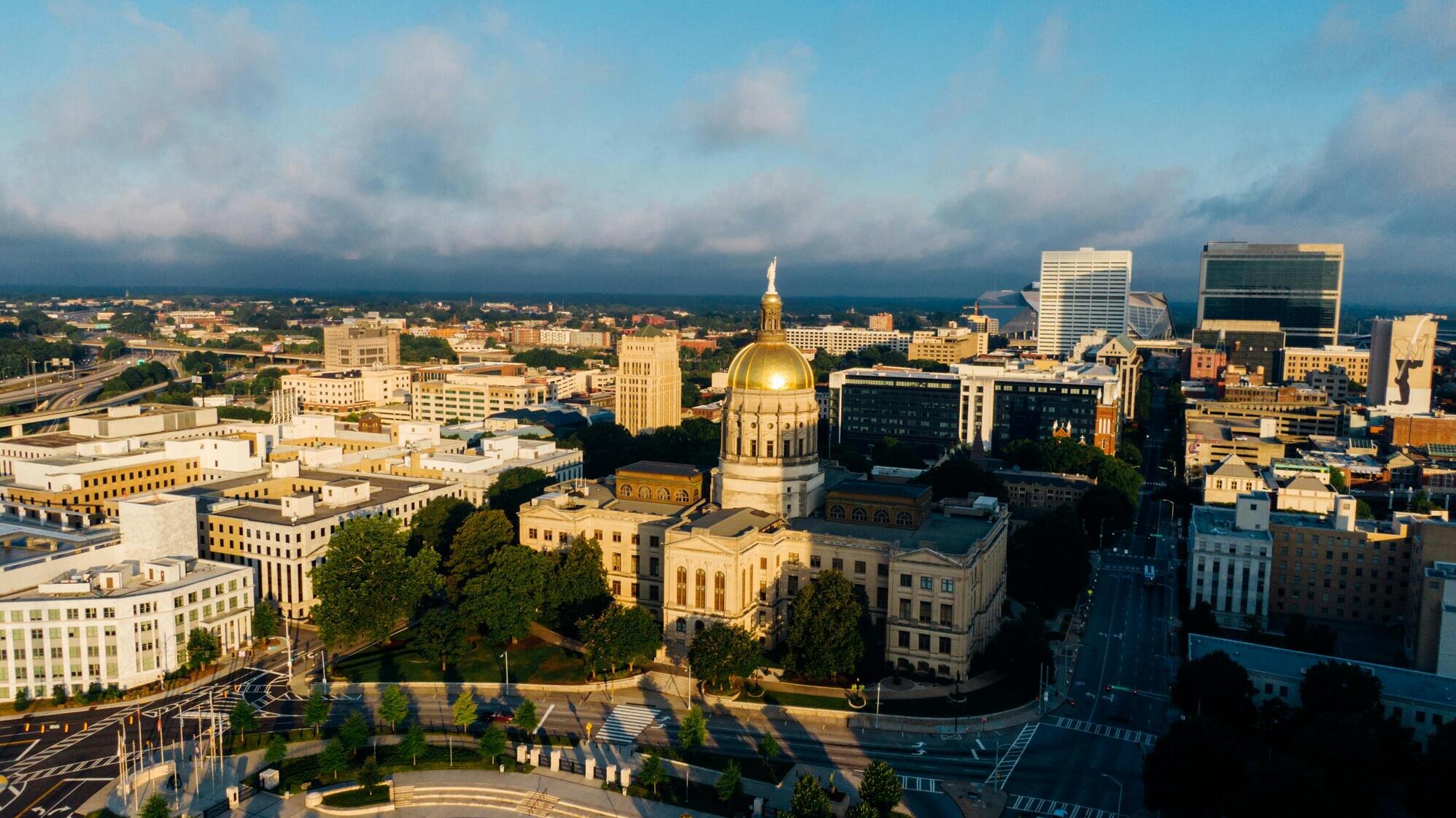 Aerial view of a city with a prominent golden-domed building surrounded by trees, roads, and modern architecture under a partly cloudy sky.