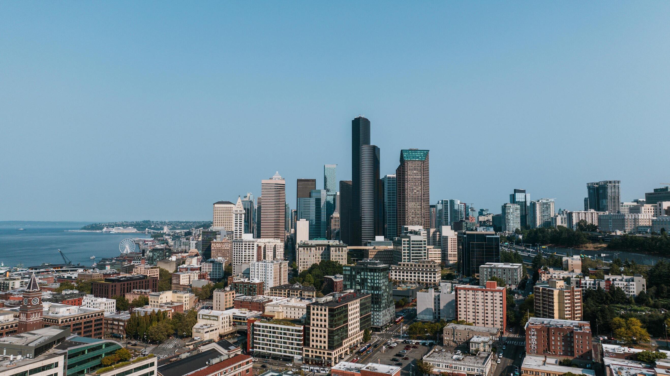 A city skyline featuring tall modern buildings, a Ferris wheel by the waterfront, and a clear blue sky.