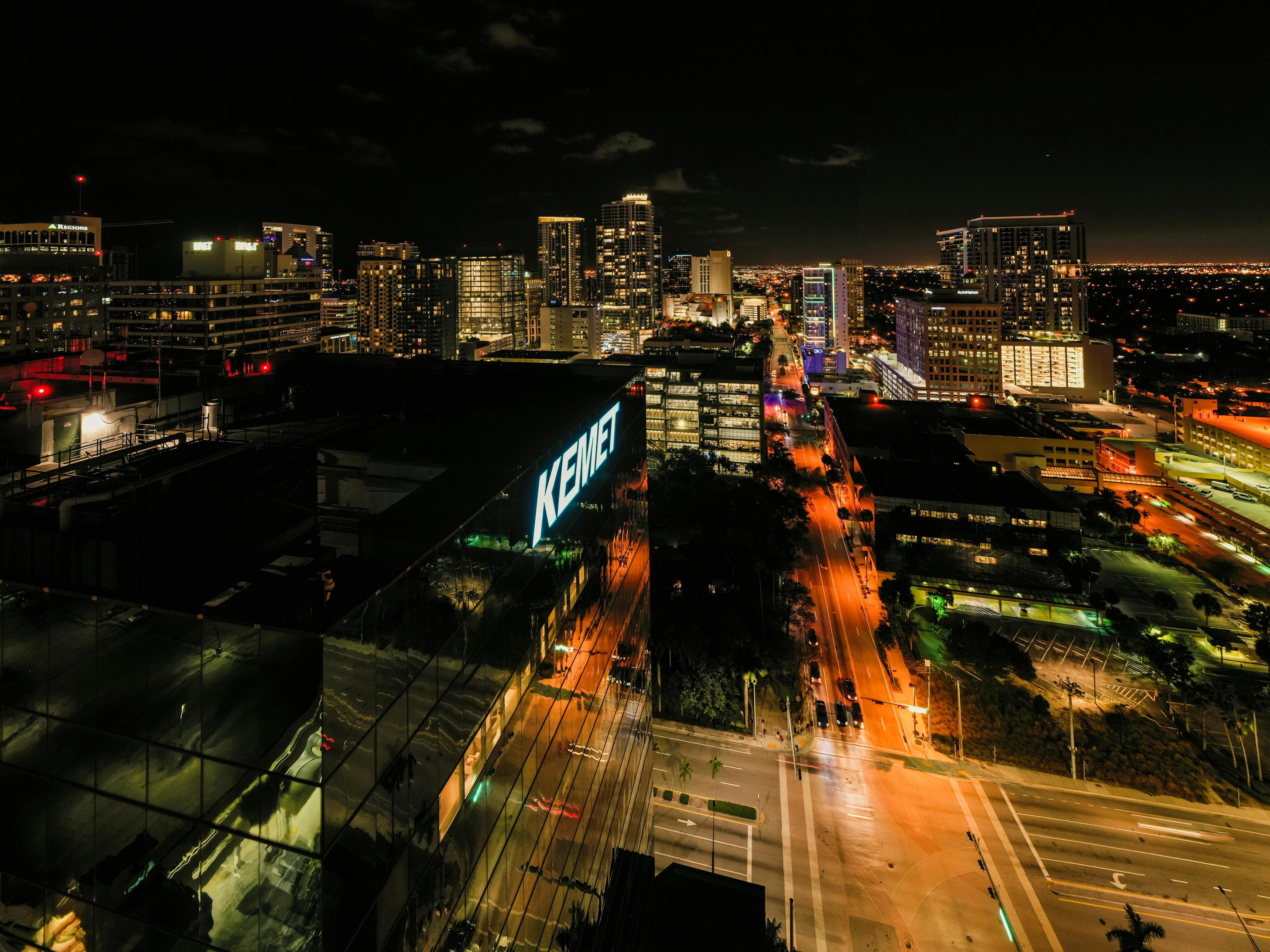 Nighttime cityscape featuring illuminated buildings, streets with glowing traffic lights, and the "KEMET" sign prominently visible on a modern structure.