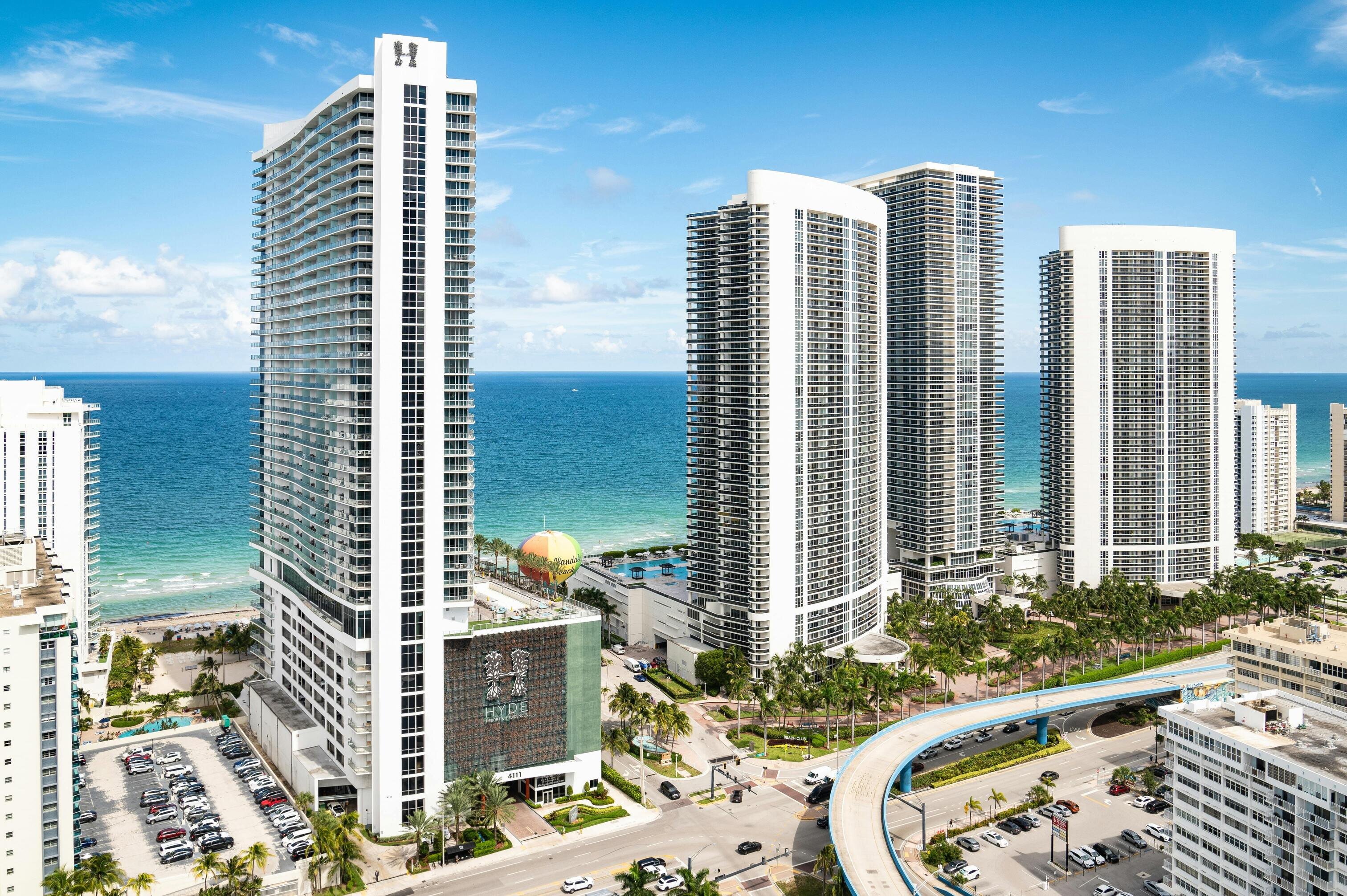 Aerial view of modern high-rise buildings by the ocean, featuring a curved road and palm trees under a clear blue sky.