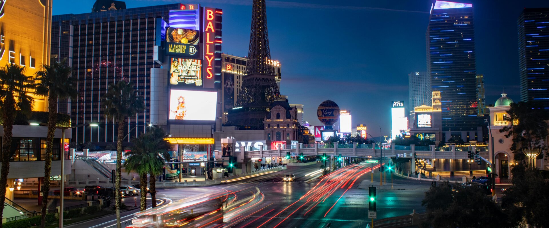 Las Vegas Strip at night with the illuminated Eiffel Tower replica and Bally's Hotel, showcasing vibrant city lights and busy traffic trails.