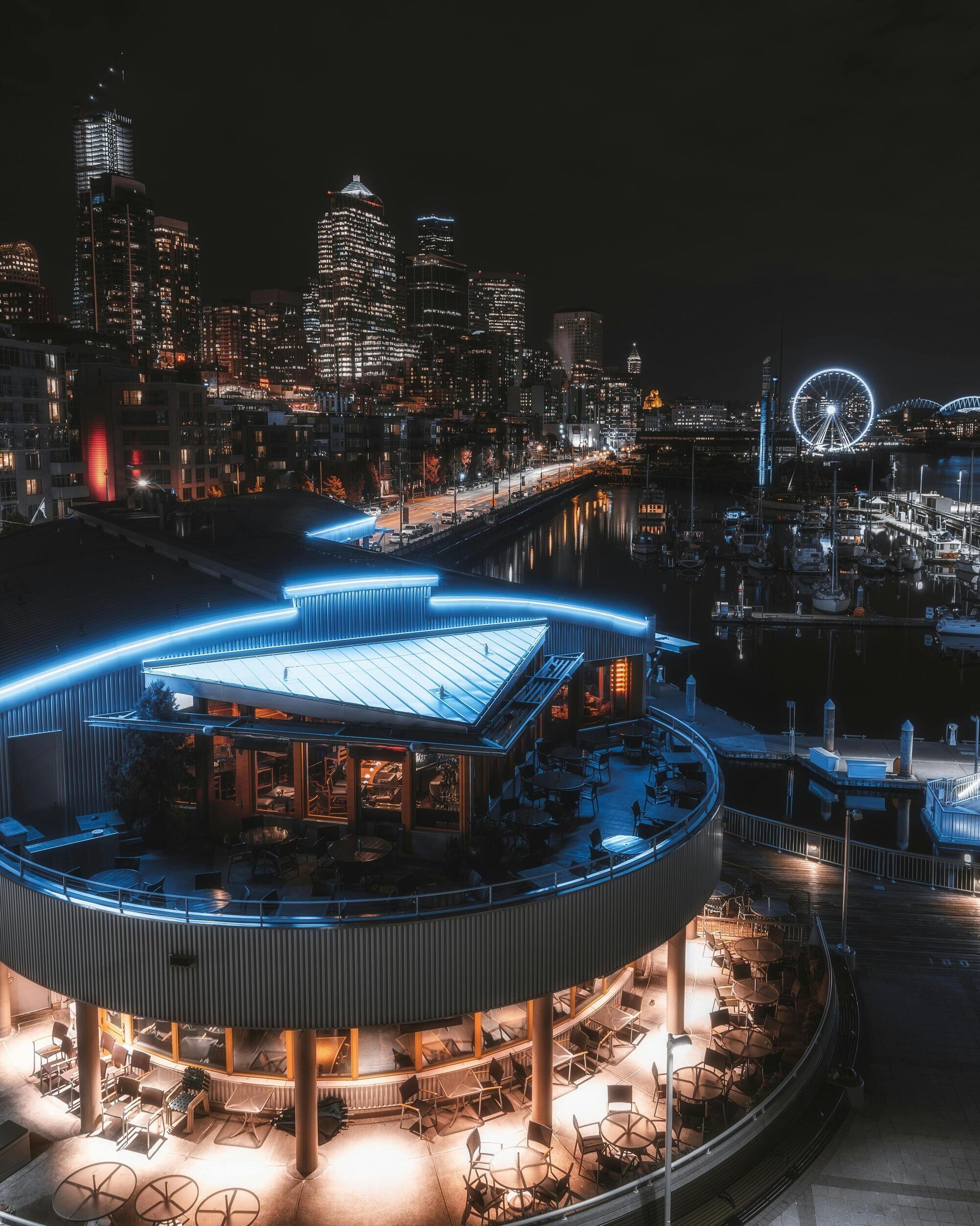A nighttime city view with illuminated skyscrapers, a Ferris wheel, and a waterfront restaurant glowing with blue and warm lights.