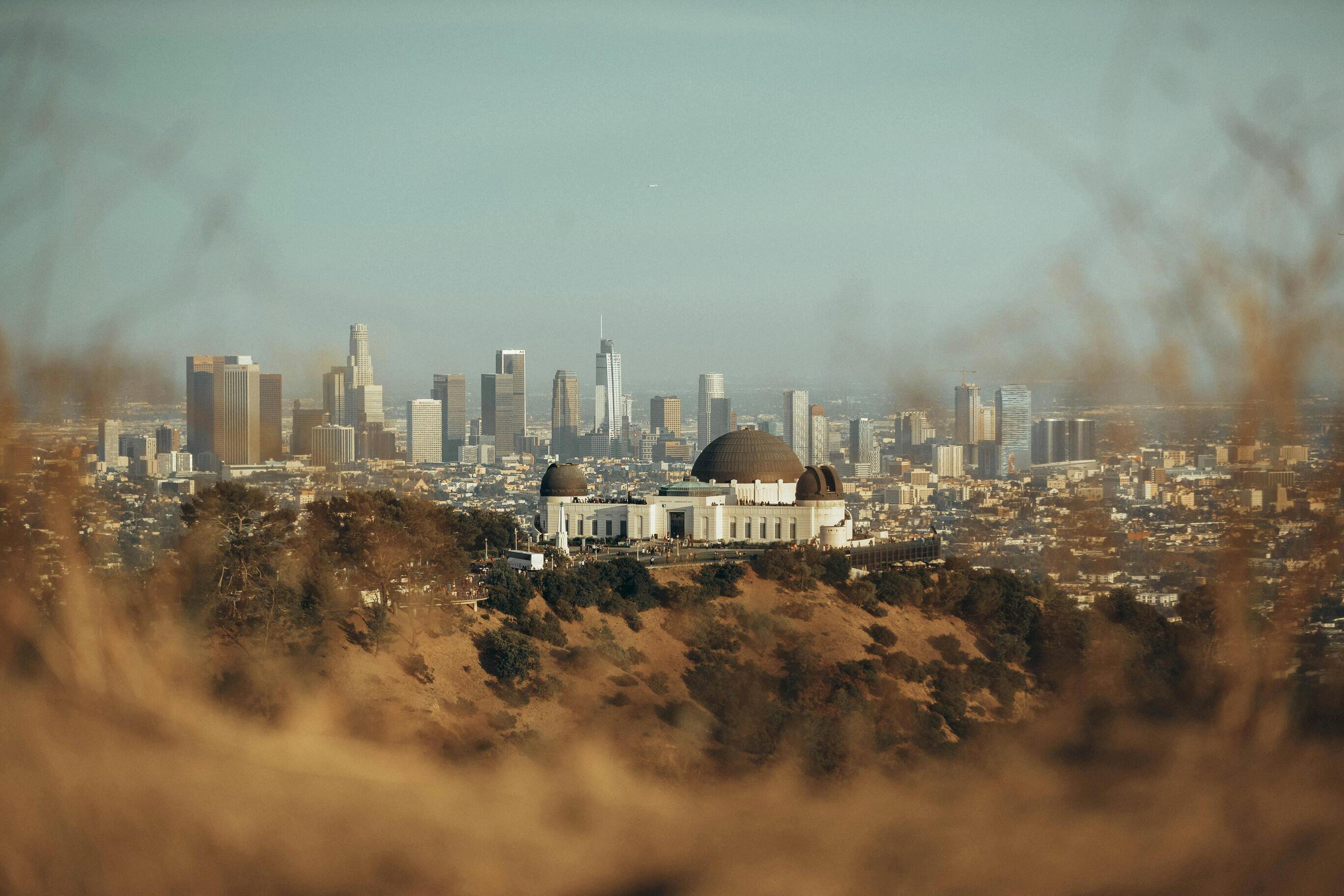 The Griffith Observatory on a hill surrounded by trees, overlooking the sprawling cityscape of downtown Los Angeles with modern skyscrapers in the background under a clear, hazy sky.