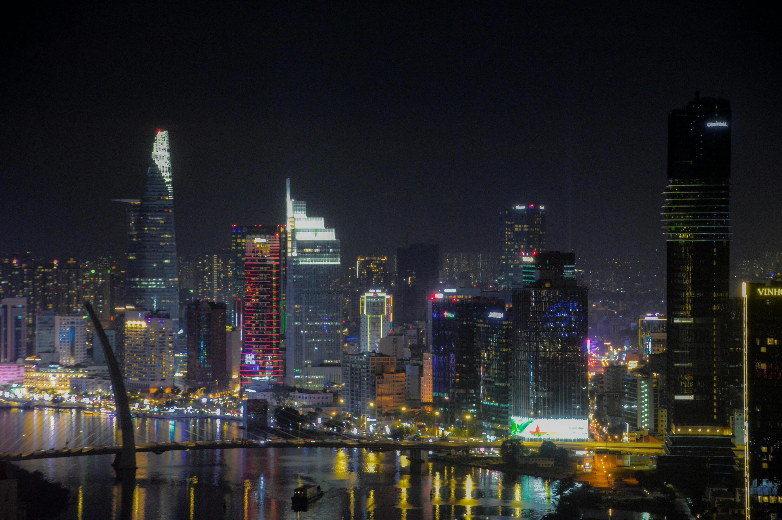 A vibrant city skyline at night with illuminated skyscrapers, colorful lights, and reflections on a river below.