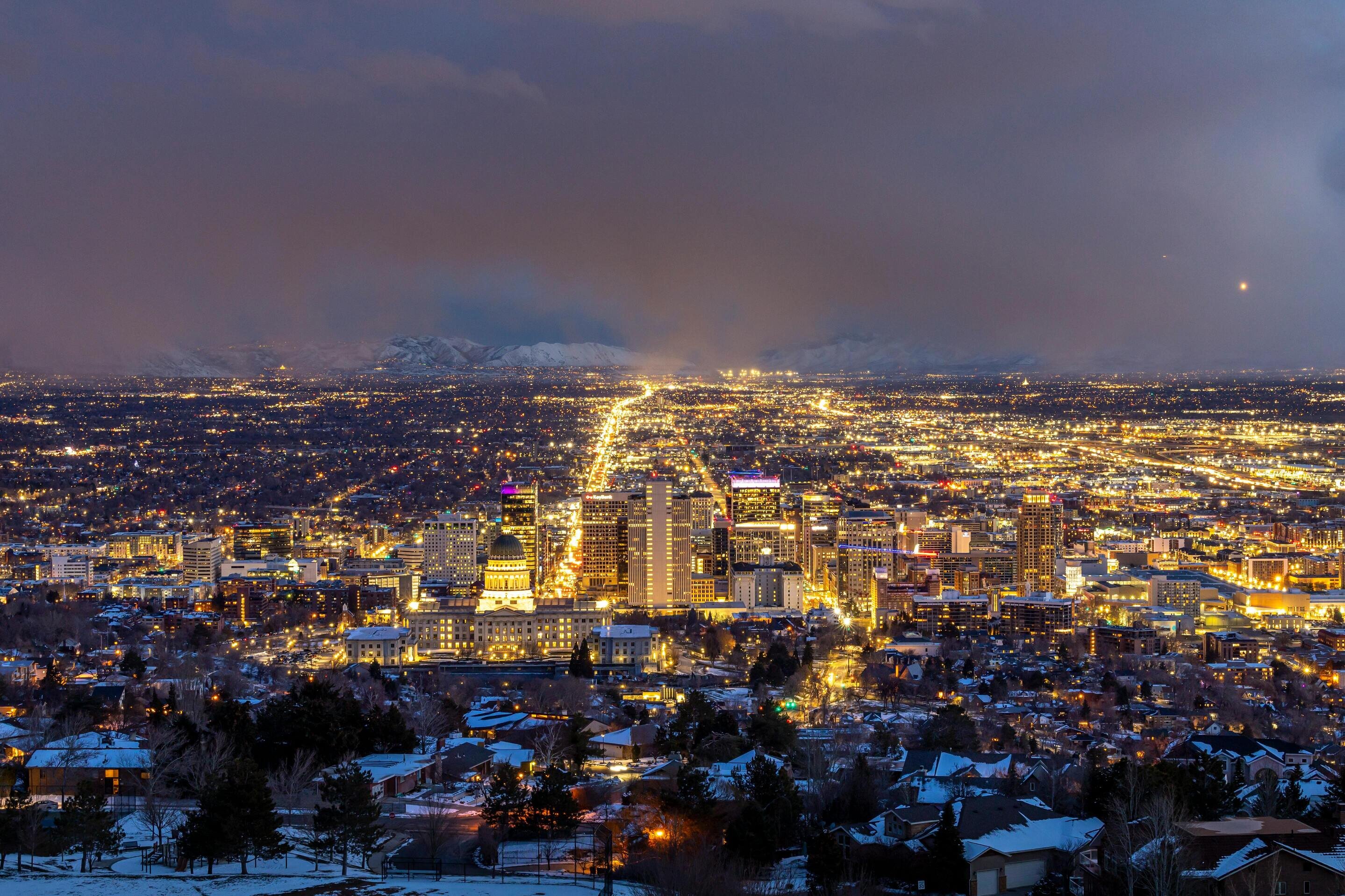 A glowing cityscape at dusk with illuminated buildings, snow-covered rooftops, and mountains in the background.