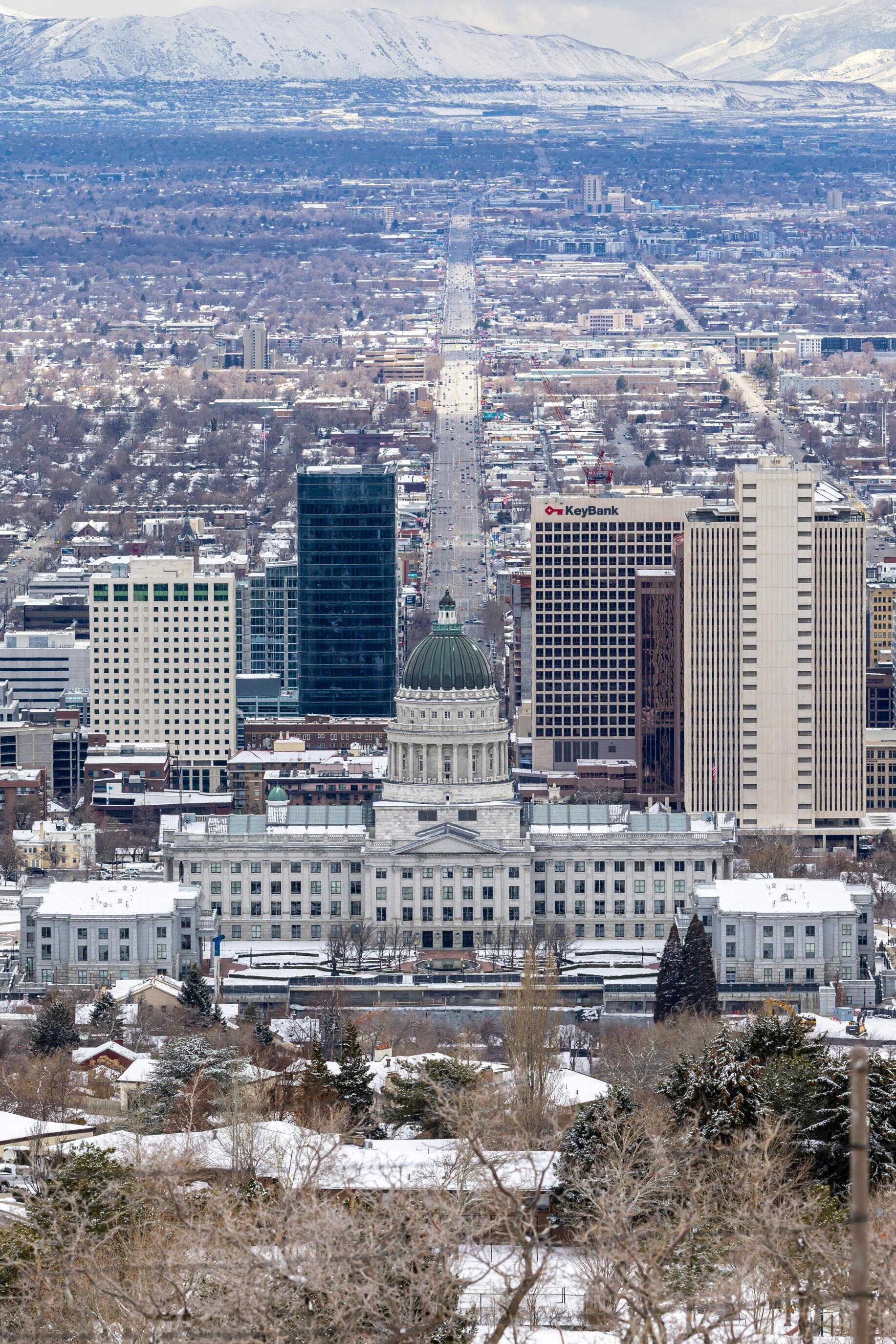 A view of the Utah State Capitol building surrounded by snow-covered rooftops, modern skyscrapers, and mountains in the distance.