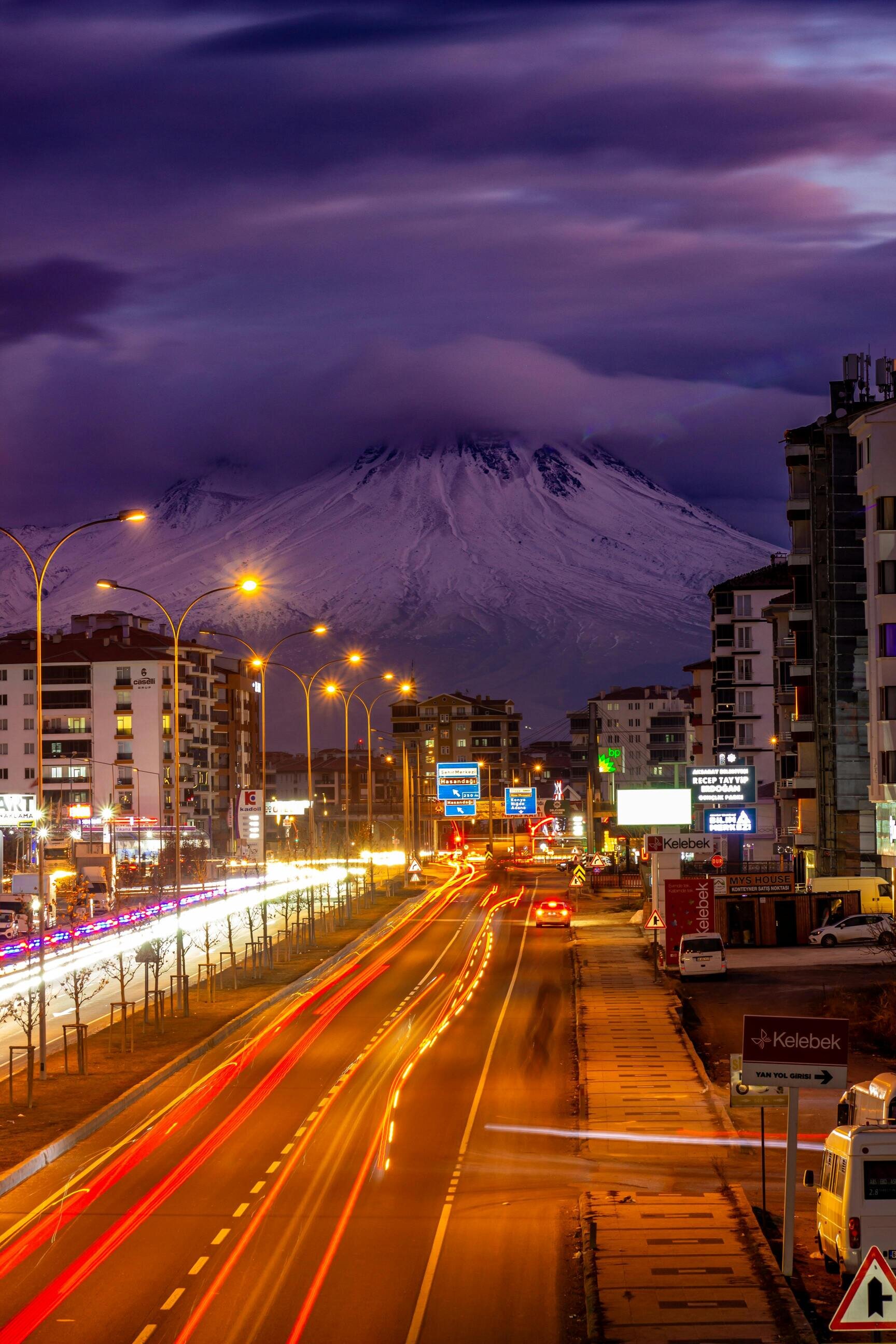 A vibrant city street at night with light trails from traffic, surrounded by buildings, and a snow-capped mountain in the background under a cloudy sky.