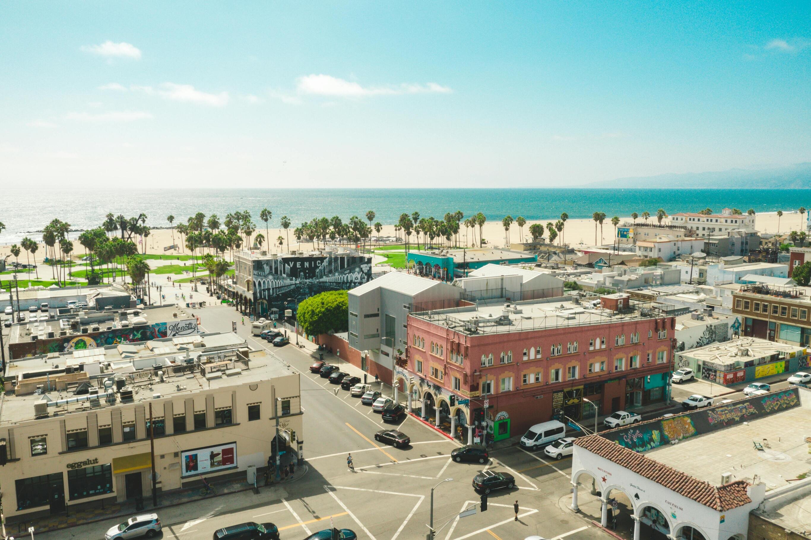 A sunny coastal neighborhood with colorful buildings, palm trees, and a sandy beach leading to the ocean under a clear blue sky.