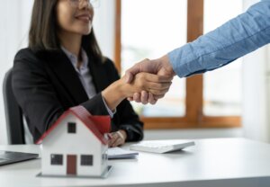 Real estate agent shaking hands with a client at a desk, with a small house model in the foreground.