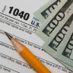 A U.S. tax Form 1040, indicating the section for personal information, is partially covered by several $100 bills, symbolizing the financial considerations and tax implications involved in selling a property.