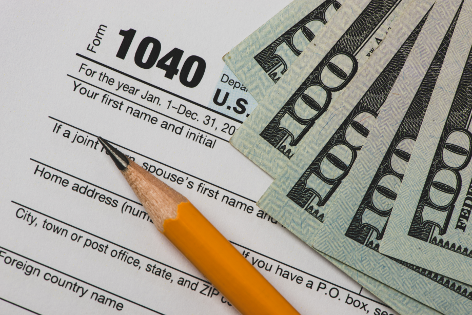 A U.S. tax Form 1040, indicating the section for personal information, is partially covered by several $100 bills, symbolizing the financial considerations and tax implications involved in selling a property.