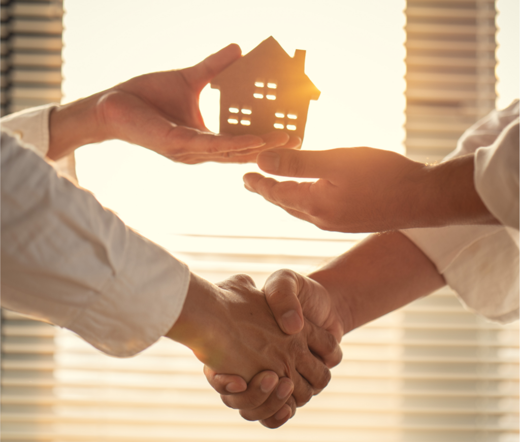 Two individuals shaking hands in a bright office setting, with one hand symbolically holding a cut-out paper house against a window, illustrating a successful cash home sale agreement between a homeowner and a real estate investor in Ohio.