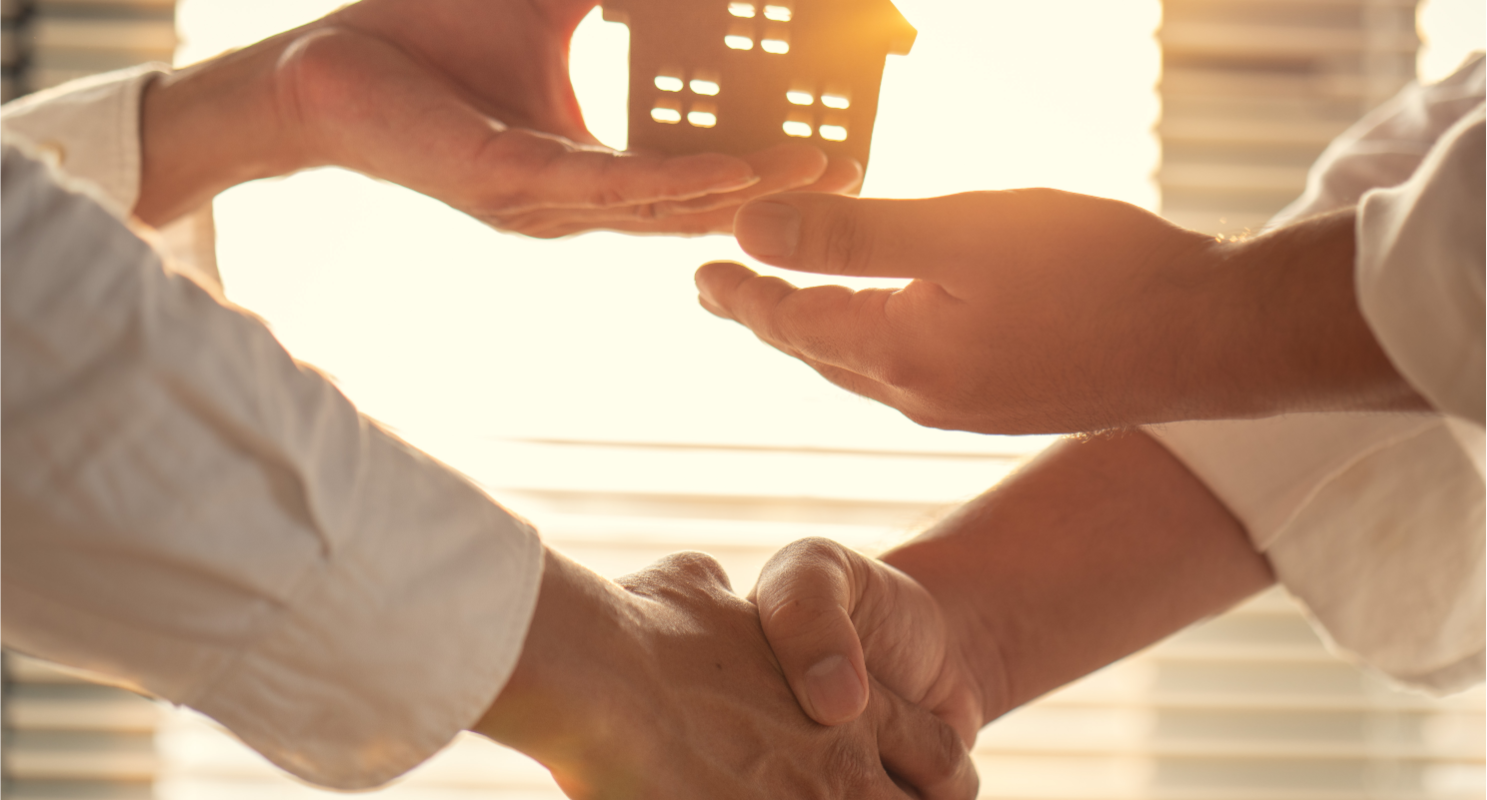 Two individuals shaking hands in a bright office setting, with one hand symbolically holding a cut-out paper house against a window, illustrating a successful cash home sale agreement between a homeowner and a real estate investor in Ohio.