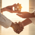 Two individuals shaking hands in a bright office setting, with one hand symbolically holding a cut-out paper house against a window, illustrating a successful cash home sale agreement between a homeowner and a real estate investor in Ohio.