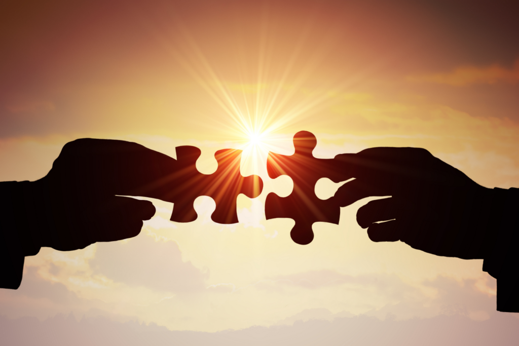 Two silhouetted hands about to connect puzzle pieces against a radiant sunset, symbolizing the perfect fit found when Ohio homeowners partner with legitimate cash-for-houses companies, culminating in a seamless and secure real estate transaction.