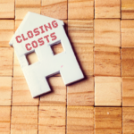 Image of a wooden block surface with a white house-shaped cutout labeled 'Closing Costs,' representing the concept of real estate closing costs in Ohio. EZ Sell Homebuyers logo in the corner.