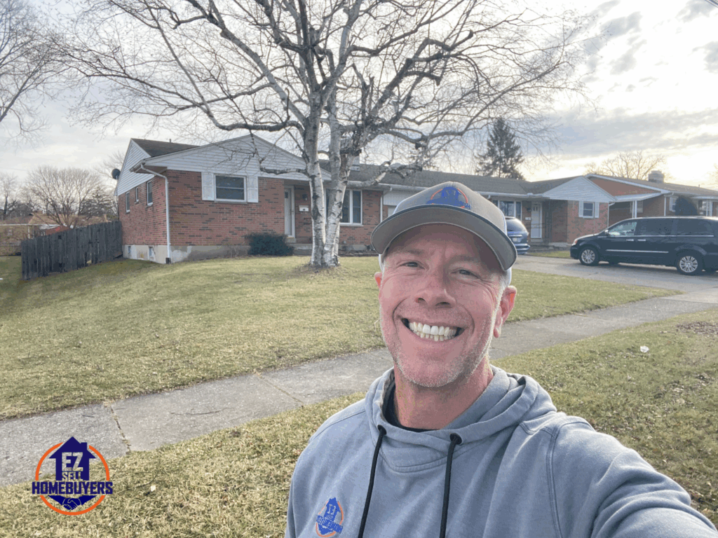 Mike Wall wearing a gray EZ Sell Homebuyers hoodie standing in front of a brick ranch home in a Dayton OH suburban neighborhood during late fall.
