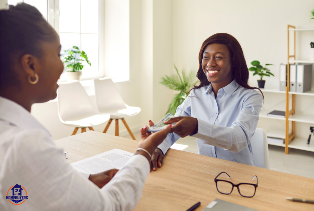 Smiling woman handing cash to another person across a desk in a bright office setting, symbolizing the process of borrowing against an inherited house in Ohio. Office features light decor, green plants, and documents on the table, reflecting a professional and approachable financial environment. EZ Sell Homebuyers logo visible in the corner, emphasizing real estate solutions.