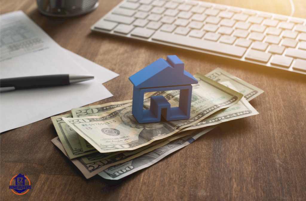 Stack of dollar bills with a blue house model on a wooden desk, symbolizing the financial benefits of seller financing in Ohio; keyboard and documents in the background.