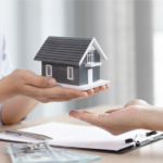 A person handing over a model house during a real estate transaction with contract paperwork and cash visible.