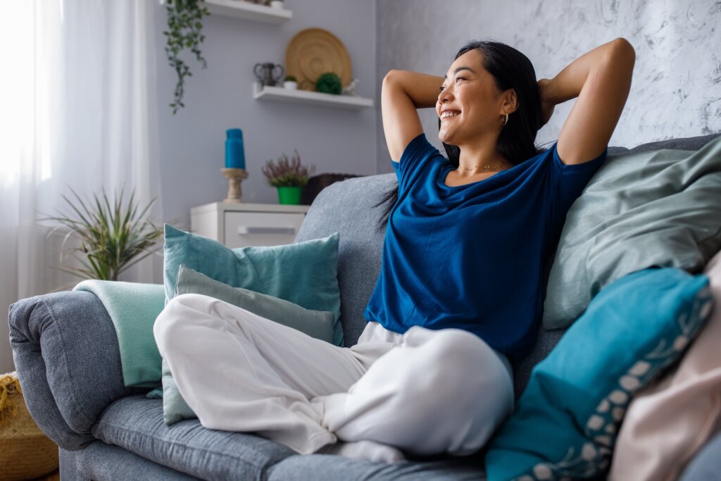 Happy young woman sitting on the cozy sofa with hands behind head, relaxed.