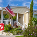 Mobile home with a 'For Sale' sign, surrounded by greenery and an American flag on the porch, showcasing a well-maintained property ready for sale.