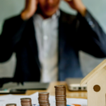 Stressed businessman sitting at a desk with a miniature house and stacks of coins, representing financial concerns and mortgage debt