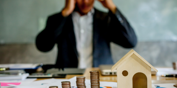 Stressed businessman sitting at a desk with a miniature house and stacks of coins, representing financial concerns and mortgage debt.