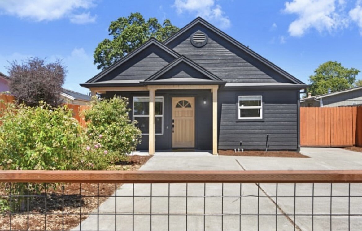 Front view of a small modern black house with a light wood door and fenced driveway on a clear day.