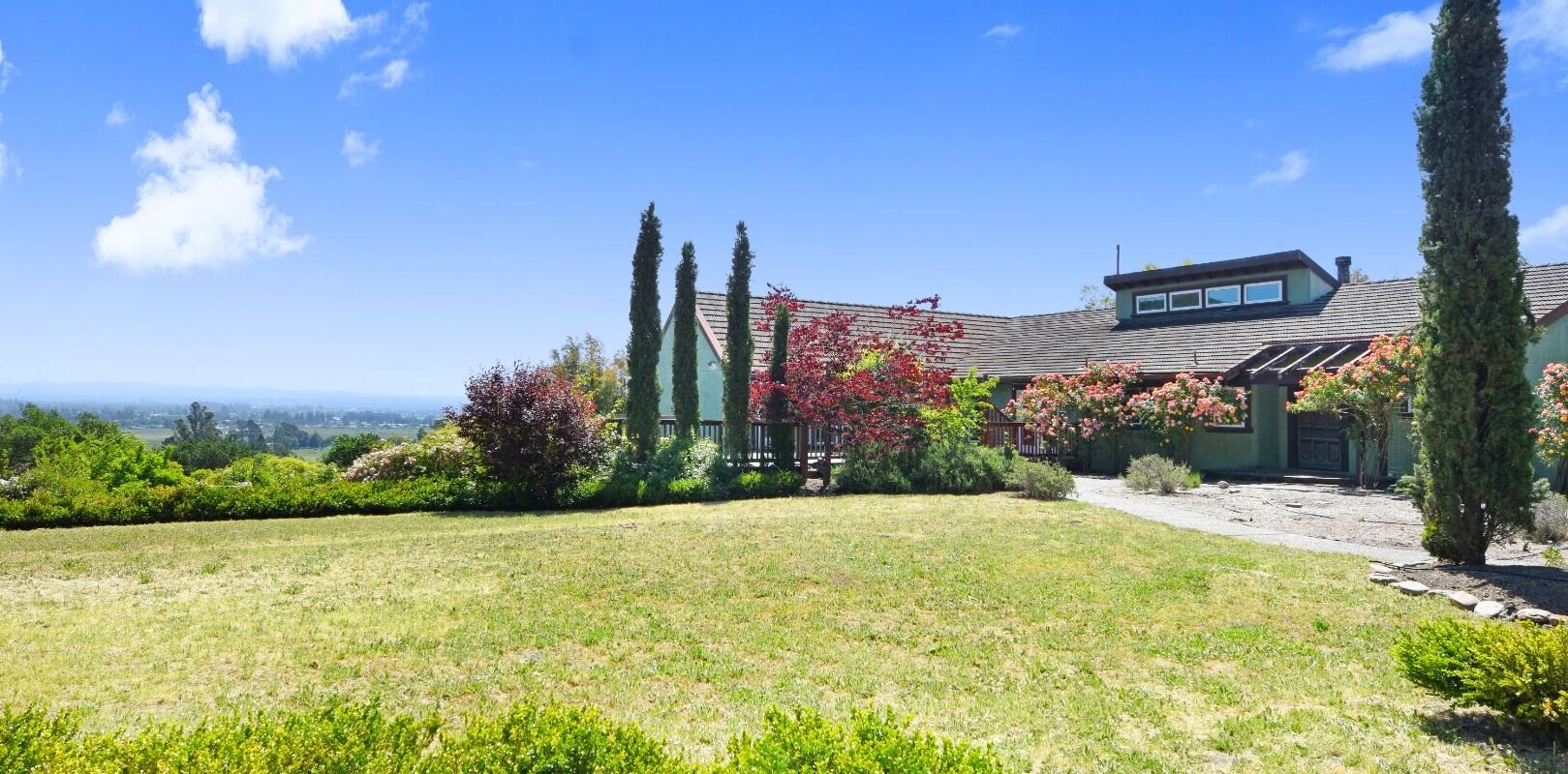 Large country home with tall cypress trees and open lawn under a bright blue sky.