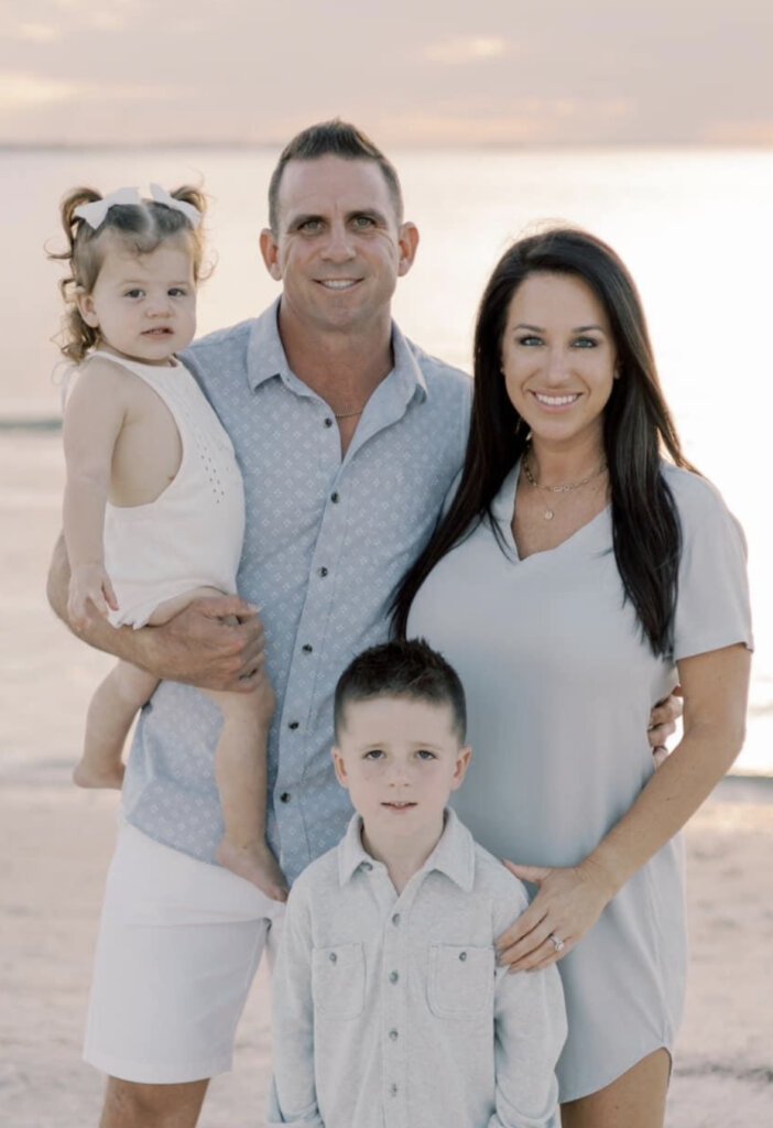 Owner of A+ Home Buyers, Mike Youngblood posing for a photo with his family on the beach. He is holding his daughter, Capri and has his arm around his wife, Gina. His son luca is standing in front of them. Everyone is smiling. Very happy family.