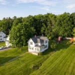 Aerial view of a white country home on a large lawn surrounded by trees, illustrating key steps to sell a rural house with Title V septic compliance.