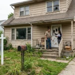 A real estate agent discusses options with a homeowner on the porch of a rundown house needing major deferred repairs, beside a yard sign marked 'Sold As-Is', representing how to sell a fixer upper in Plymouth, MA.