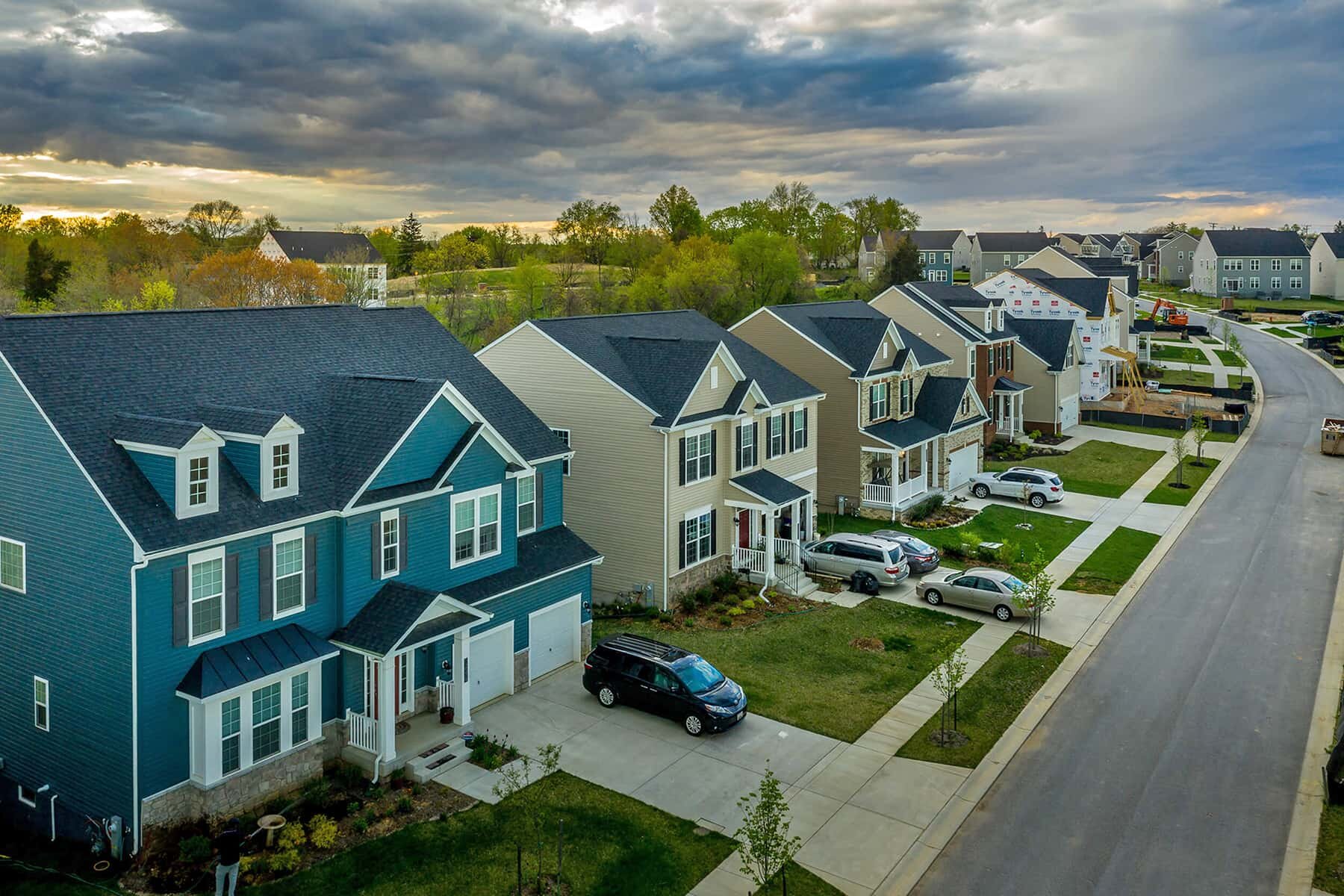 Aerial view of a suburban neighborhood with rows of modern two-story homes, well-maintained lawns, and cars parked in driveways, under a cloudy sky at sunset.