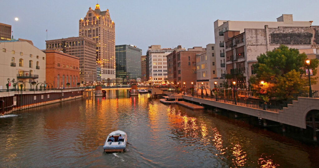 Evening view of a city riverfront, featuring illuminated buildings and a boat cruising on the water, ideal for urban real estate settings.