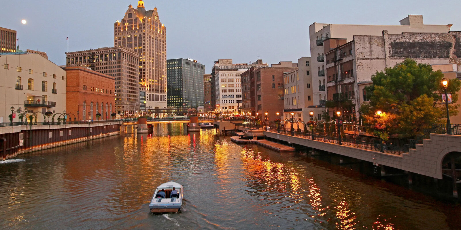 Evening view of a city riverfront, featuring illuminated buildings and a boat cruising on the water, ideal for urban real estate settings.