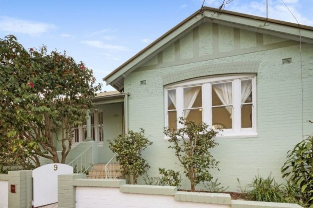 A charming mint green house with a gabled roof, arched windows, and a small front garden, surrounded by trees and plants.