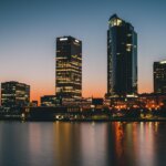 A stunning city skyline at dusk, featuring a variety of illuminated high-rise buildings reflecting on a calm water surface. The sky transitions from a deep blue at the top to a vibrant orange near the horizon, enhancing the silhouette of an iconic, sail-like structure to the left. This serene evening view captures the essence of urban beauty and tranquility.