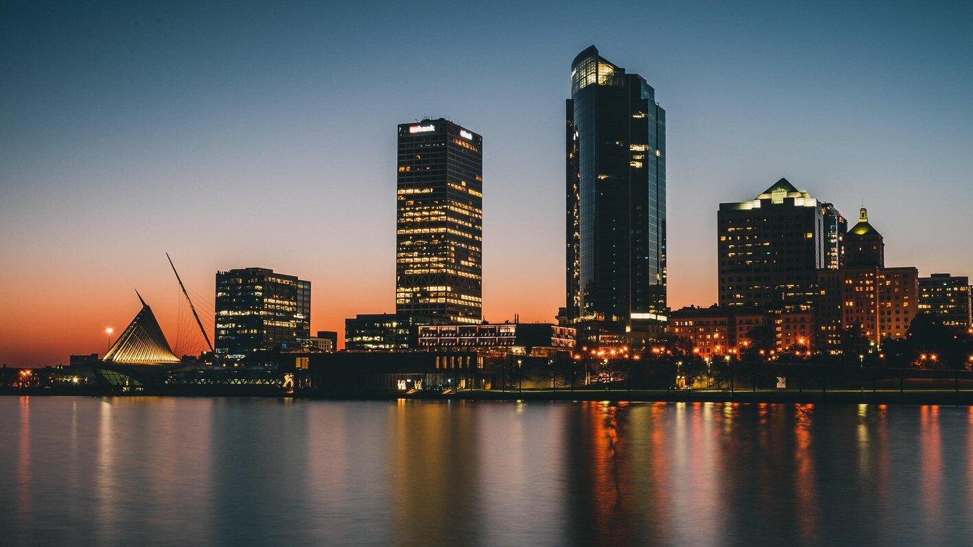 A stunning city skyline at dusk, featuring a variety of illuminated high-rise buildings reflecting on a calm water surface. The sky transitions from a deep blue at the top to a vibrant orange near the horizon, enhancing the silhouette of an iconic, sail-like structure to the left. This serene evening view captures the essence of urban beauty and tranquility.