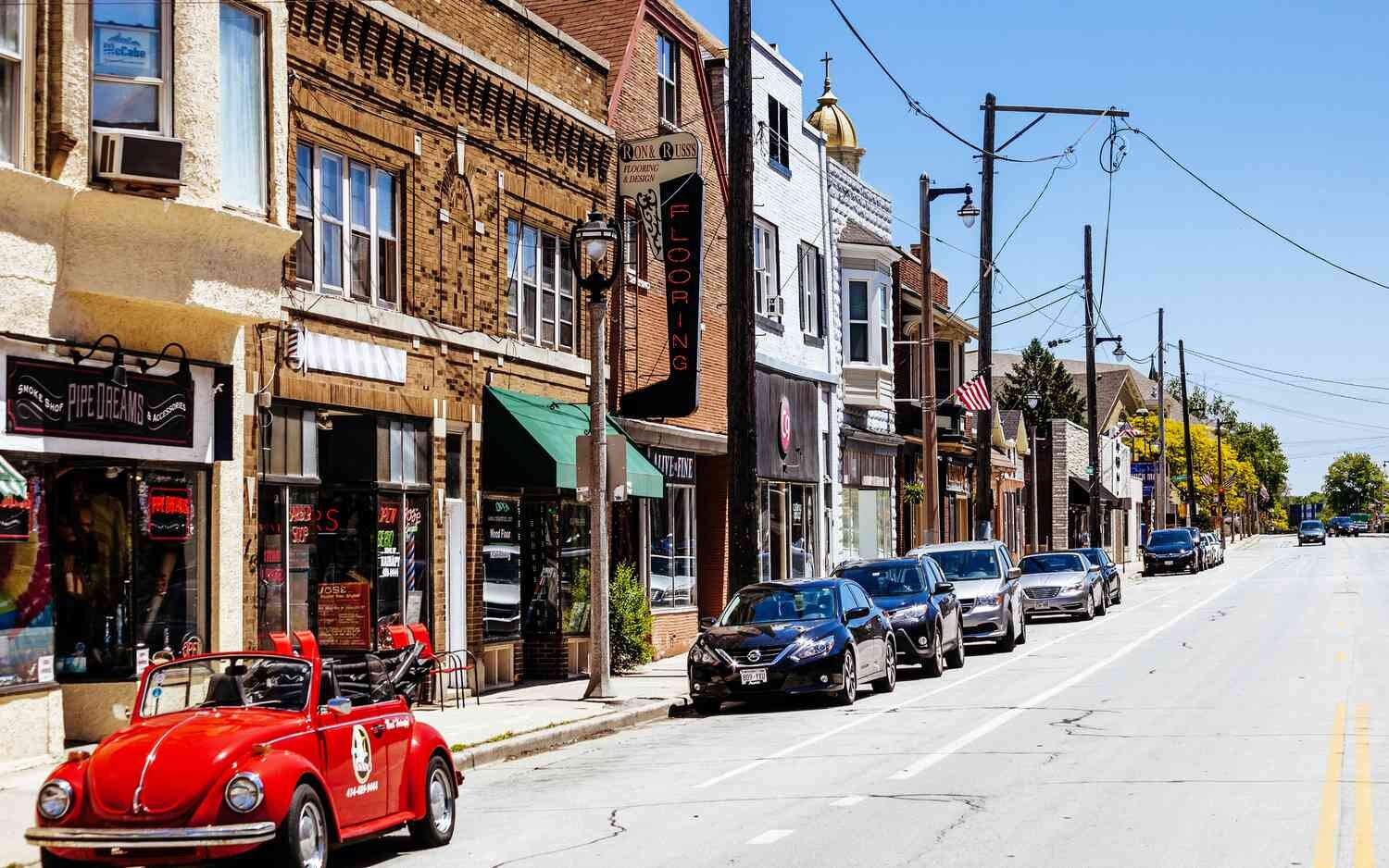 A street lined with small shops and businesses in an older neighborhood, with a mix of historic brick buildings, parked cars, and a bright red convertible in the foreground.