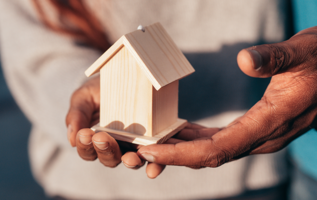 Close-up of hands gently holding a small wooden house model, symbolizing homeownership, real estate, or housing security.