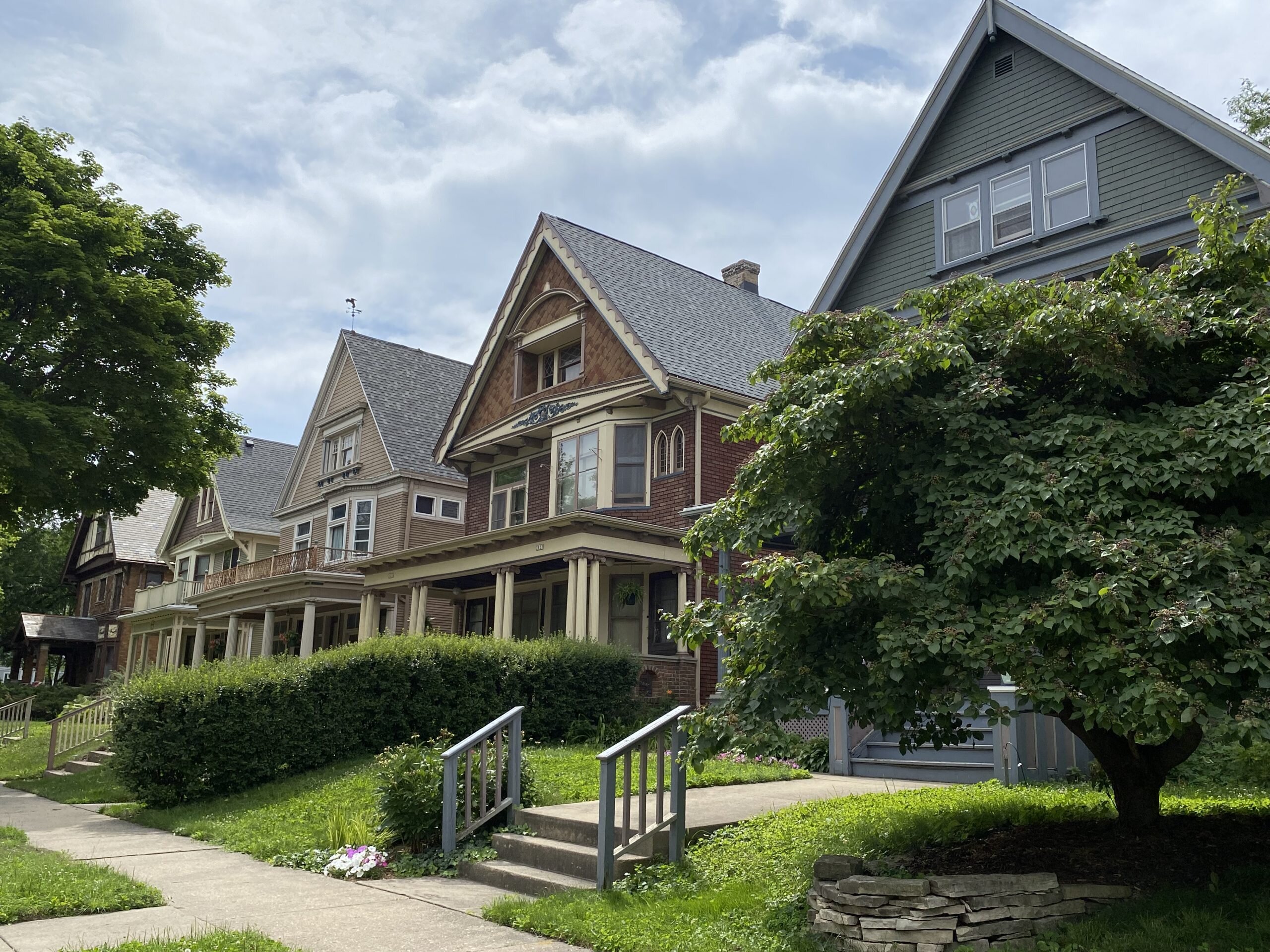 Image of a row of charming, older homes in a residential neighborhood, featuring Victorian-style architecture with gabled roofs and large front porches, surrounded by lush green trees and shrubs.