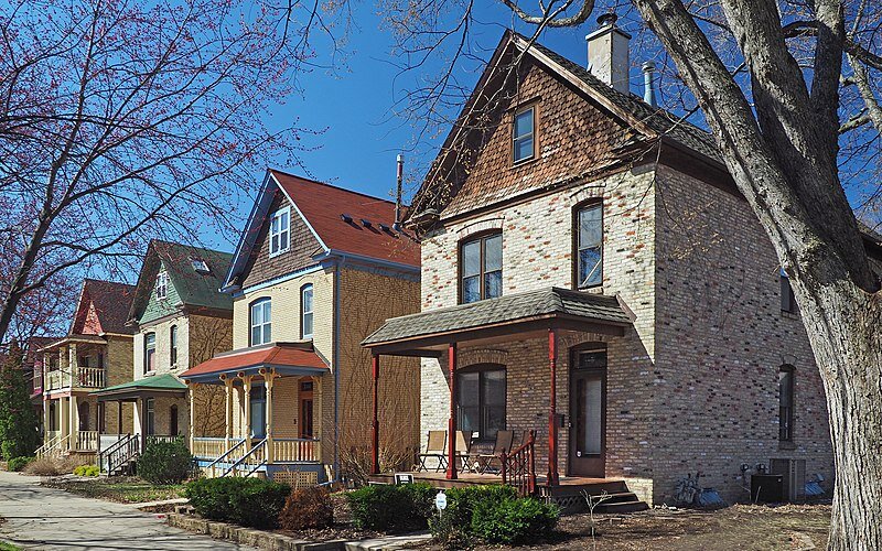 Three historic brick houses with gabled roofs and front porches on a quiet residential street, with trees suggesting early spring or late autumn.